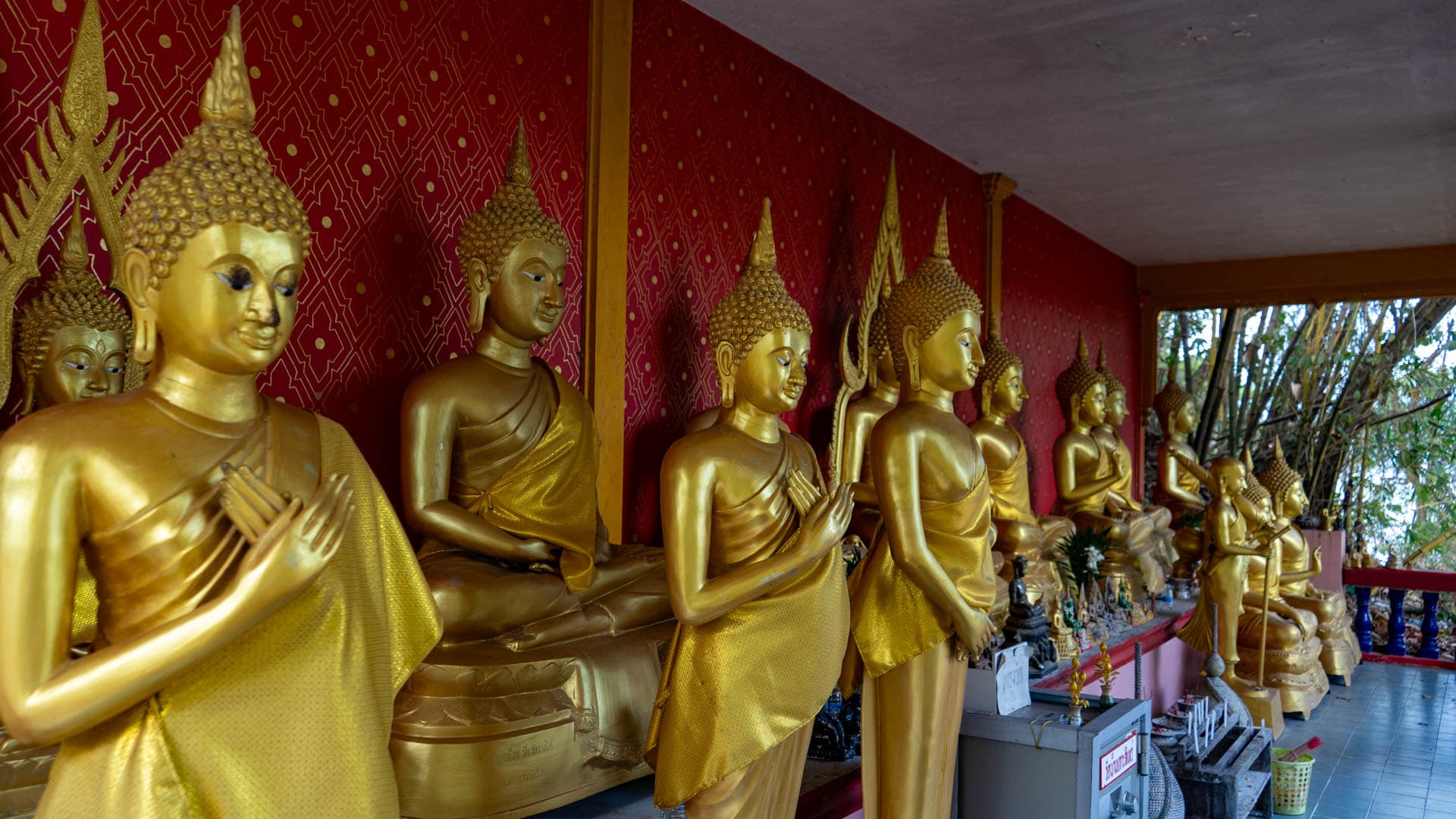 Standing Buddha statues along red shrine wall at Wat Ban Koh Sirey