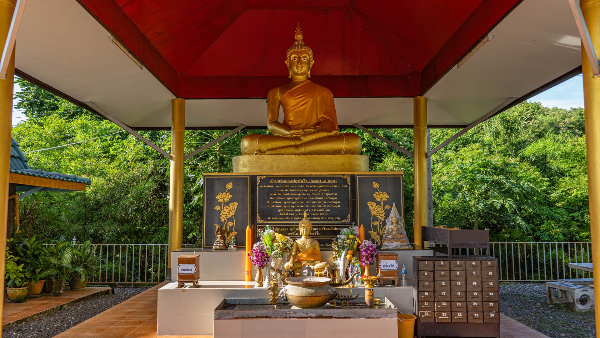 Seated Buddha in pavilion with offerings and green foliage behind