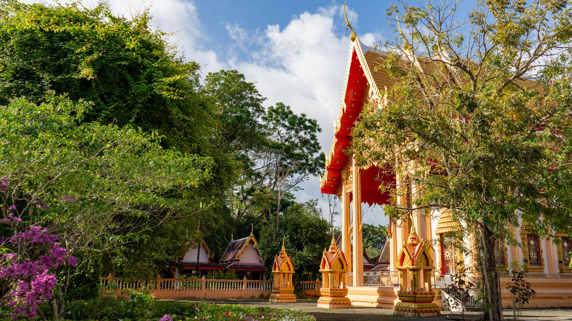 Side of Wat Phra Thong main hall with shrine pavilions, trees, and planted garden edge