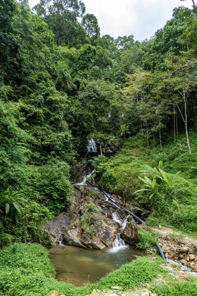 Portrait view of narrow jungle valley with multi-tier waterfall cascade over granite boulders, water pipe crossing foreground rocks, dense rainforest canopy above, Kamala Phuket