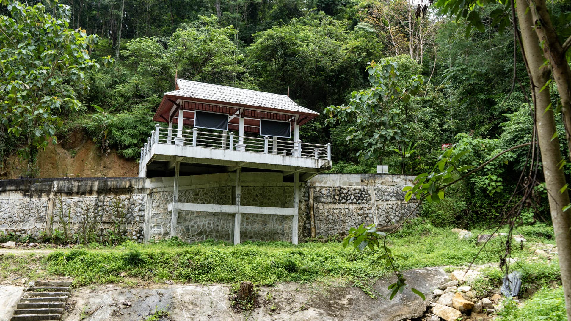 Narrow paved road curving uphill beside concrete water gate and reservoir, forested hillside behind, pickup truck passing, Kamala village visible in distance
