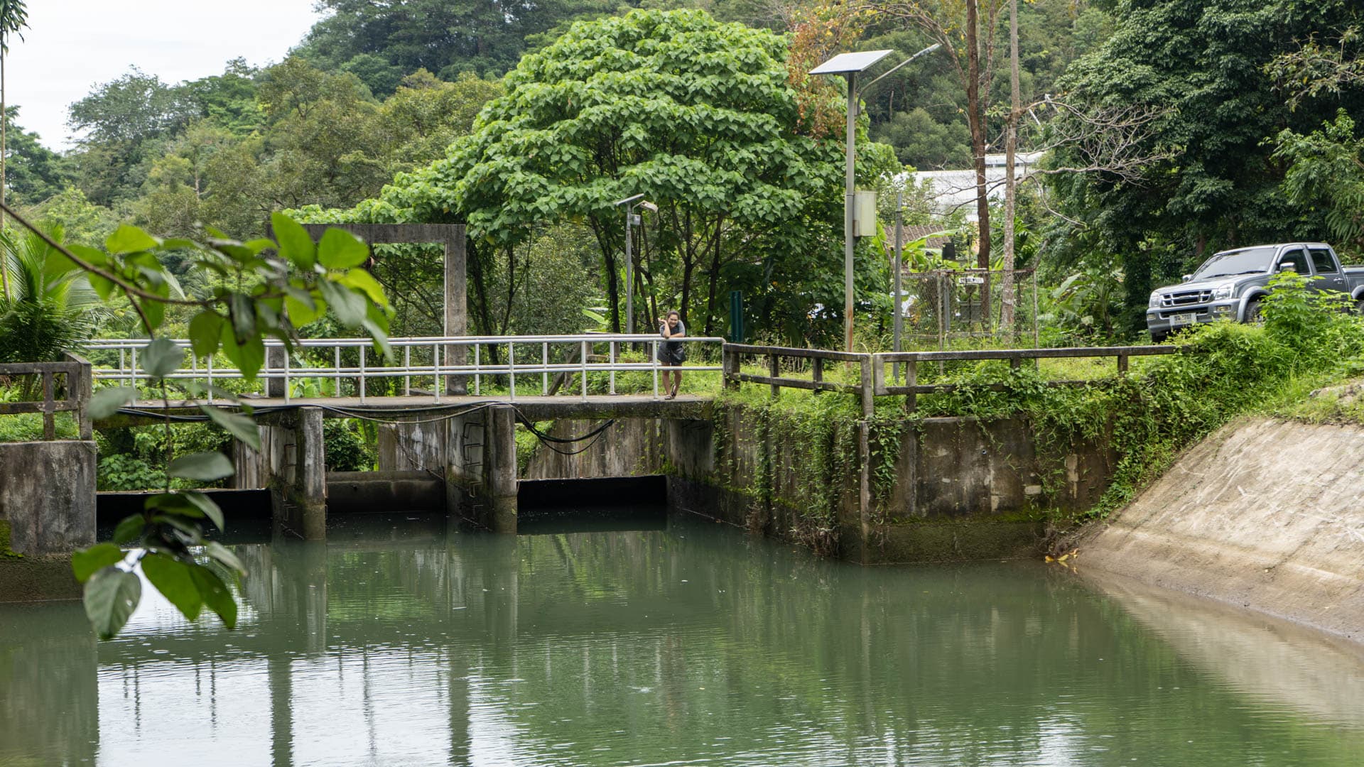 Concrete water control gate with metal railings spanning a still green reservoir, forested hill rising behind, single person standing on the crossing, pickup truck parked right