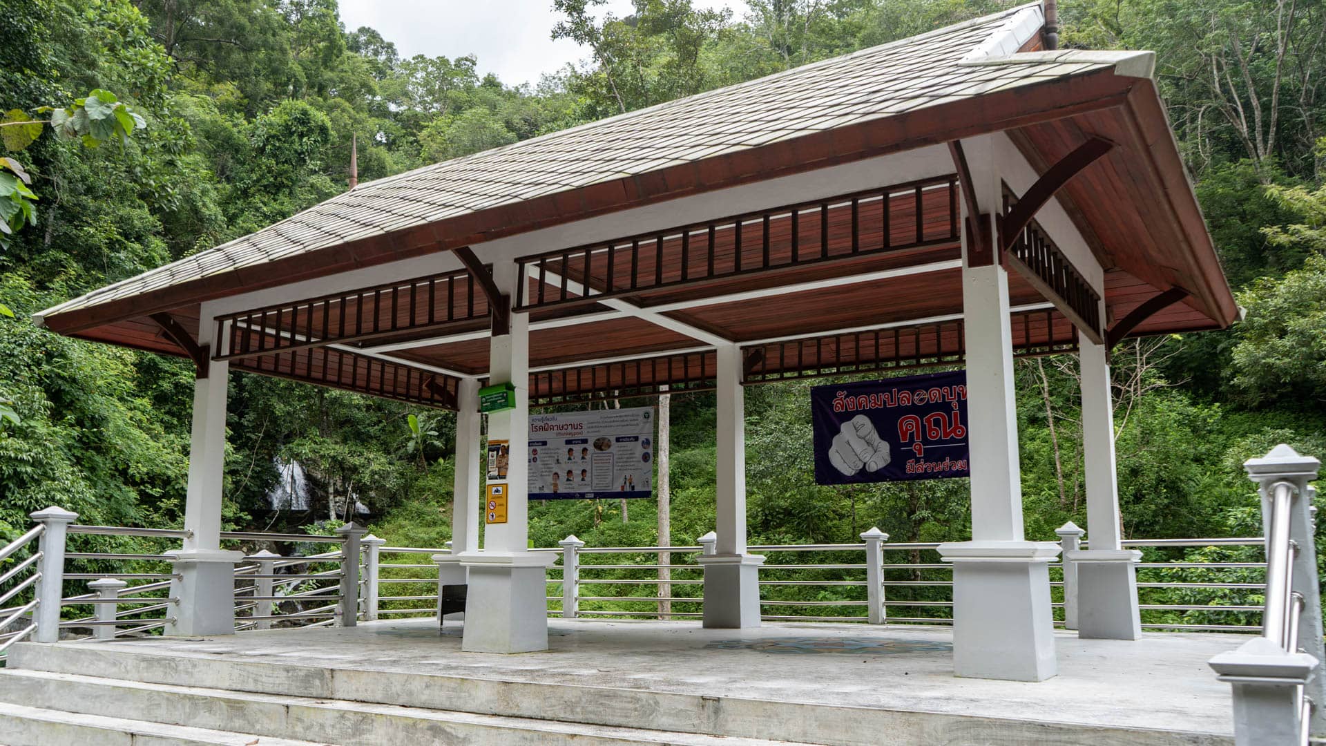 Open-sided Thai-style pavilion with timber and tile roof, concrete platform, information boards in Thai mounted inside, dense rainforest behind, waterfall visible left
