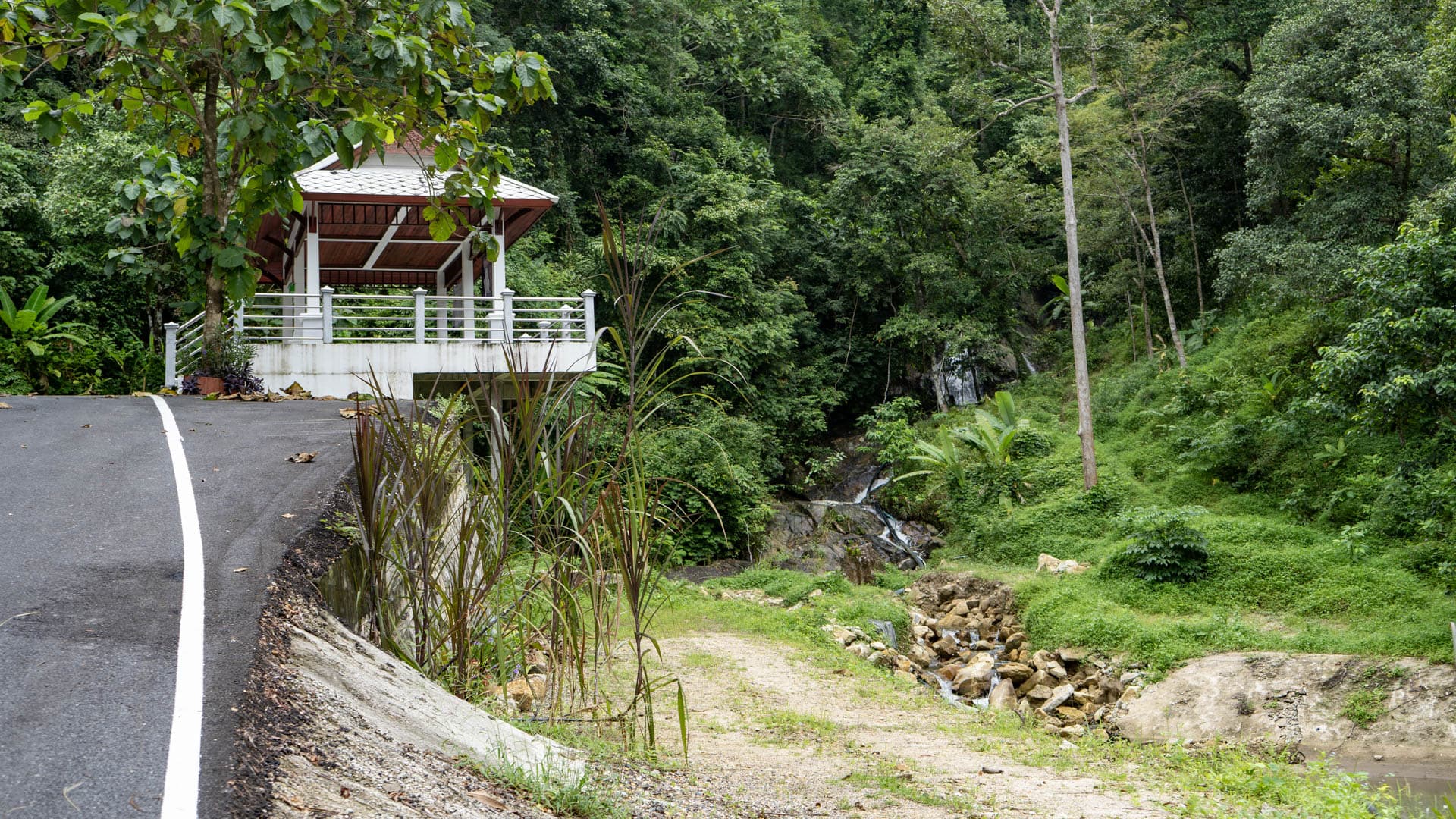 Paved road curving left past white-roofed open sala structure on stilts, waterfall cascade visible through vegetation on right, forested hillside rising steeply