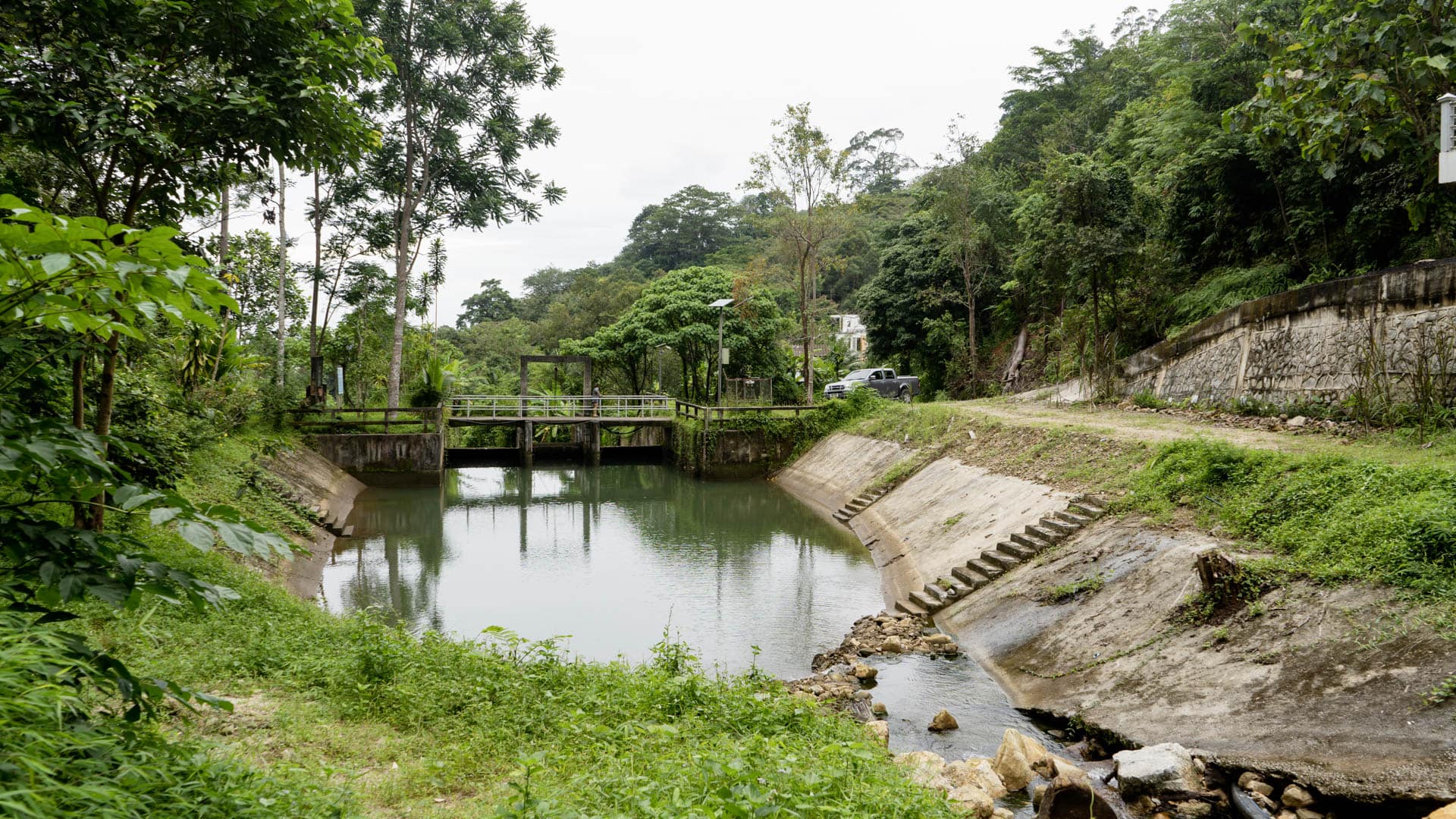 Concrete-edged reservoir with control gate bridge, stepped concrete channel to right, unpaved track and pickup truck beyond, forested hillside and building visible behind