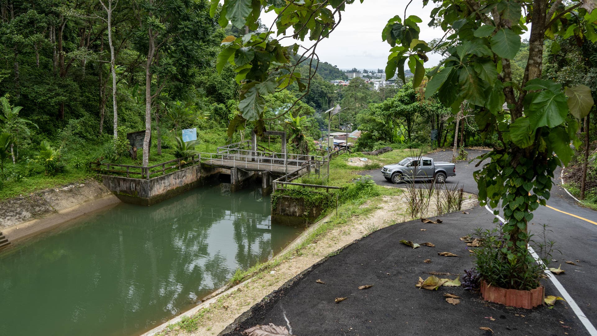Reservoir and water gate left, paved road curving right with pickup truck, vine-covered tree foreground, Kamala village buildings visible on forested hillside behind