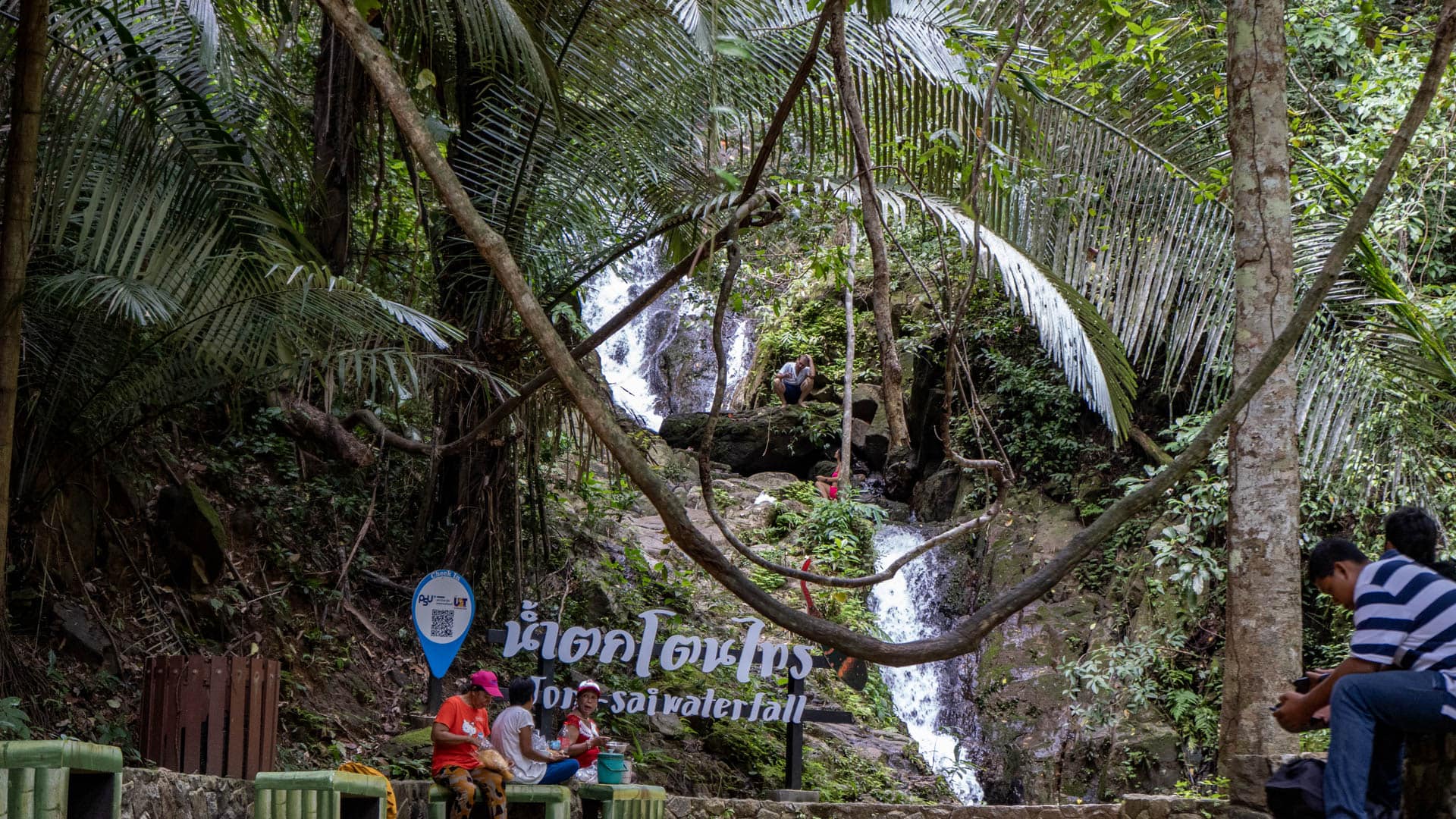 Bilingual Thai and English Ton Sai waterfall sign with visitors seated on benches, waterfall and palm fronds visible behind, Phuket