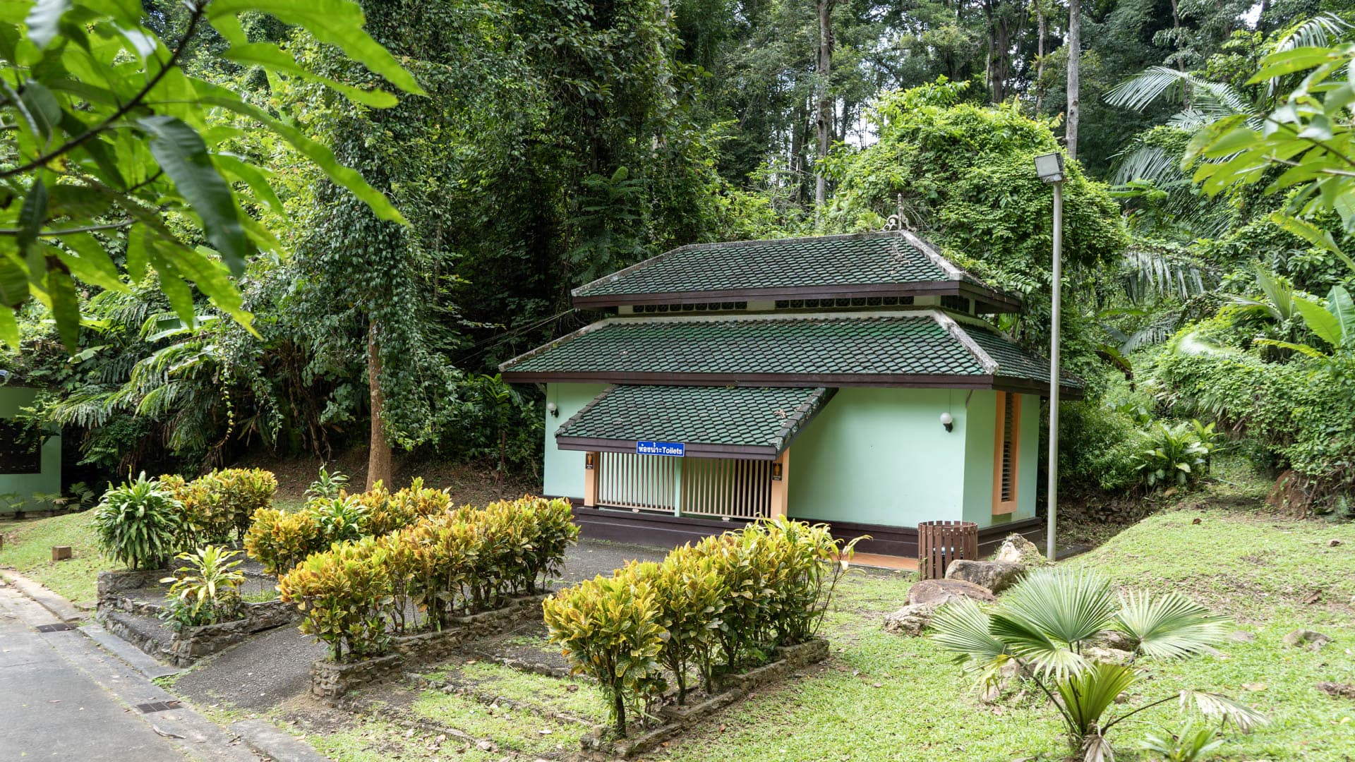 Small green-roofed toilet block with manicured garden beds set against tall rainforest canopy, paved path at left, Phuket