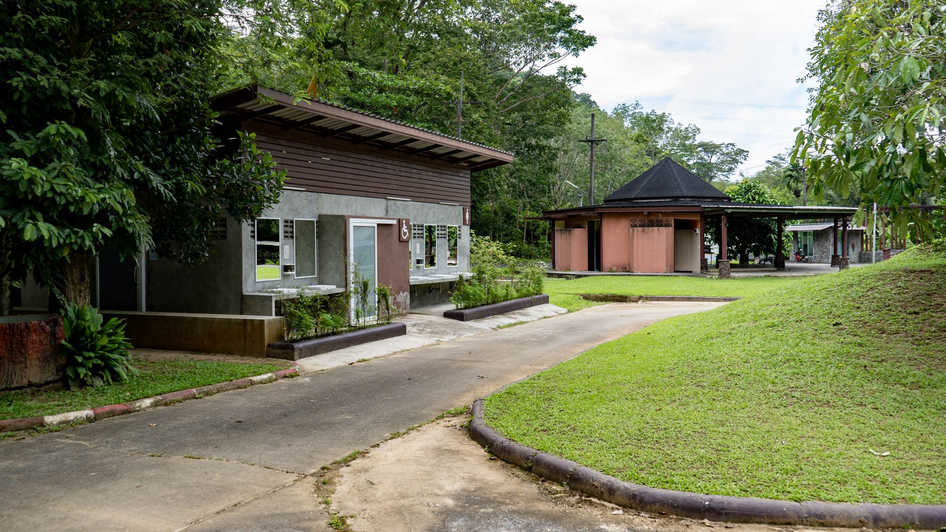 Concrete path curving past accessible toilet block and open shelter buildings on mown grass, rainforest canopy behind, no visitors, Phuket